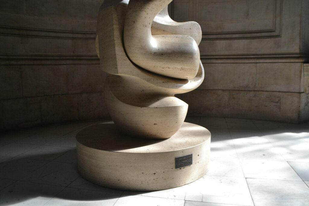  modern abstract sculpture made of stone. The sculpture features smooth, flowing curves and appears to be a stylized representation of a human figure holding a vase or vessel. The sculpture is mounted on a circular base and is set against a background of stone walls with rectangular paneling. The interplay of light and shadow on the sculpture highlights its intricate contours and textures. A small plaque on the base of the sculpture is visible, but the text on it is not legible
