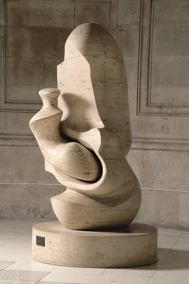 A modern abstract sculpture made of stone, placed on a circular base. The sculpture has smooth, curved surfaces and appears to be an intricate, intertwined form. There is a small plaque on the base of the sculpture with the text 'MOTHER AND CHILD HENRY MOORE.' The sculpture is situated in a dimly lit room with stone walls and a tiled floor, with light casting shadows on the sculpture and the floor. The interplay of light and shadow highlights the smooth texture and curves of the sculpture, making it visually interesting.