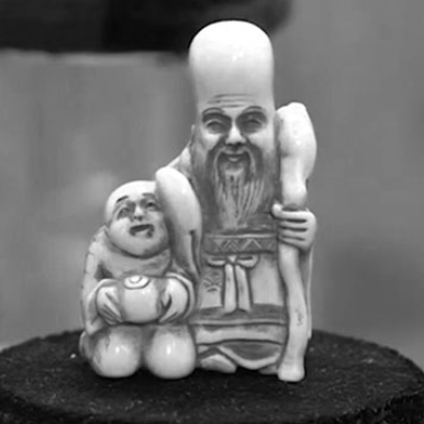A small figurine of two human-like figures. The larger figure is standing and holding a staff, while the smaller figure is kneeling and holding an object resembling a bowl or fruit. The figurine appears to be made of a light-colored material, possibly ivory or ceramic, and is placed on a dark circular platform. The background is blurred, emphasizing the figurine itself.