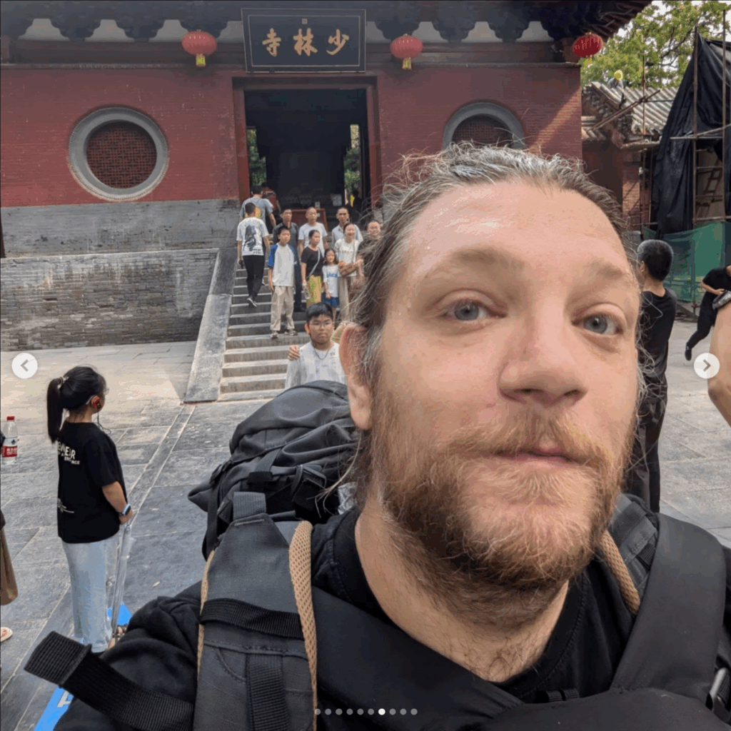 Picture of me - a bearded blonde man -with a backpack taking a selfie in front of the entrance to the Shaolin Temple in China, with a group of people posing on the steps behind him.