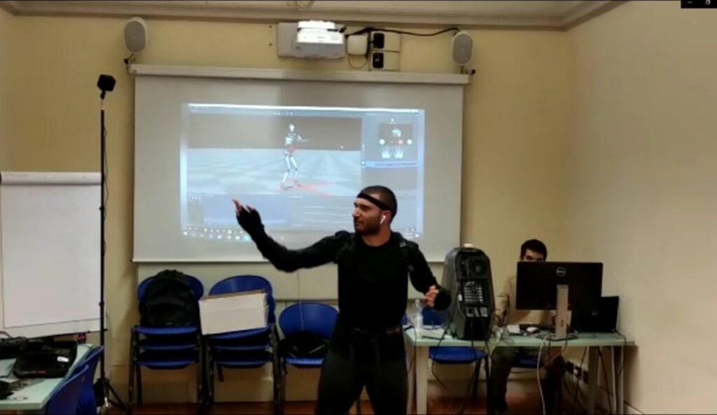 An individual wearing a motion capture suit in a room equipped with various technology for capturing and analyzing motion data. The person is standing in front of a large screen displaying a 3D model that mimics their movements. The room also features blue chairs, a whiteboard, and a tripod with a camera or sensor. Another individual is seated at a desk with a computer setup, likely monitoring or controlling the process. This setup suggests a demonstration or class focused on motion capture technology.