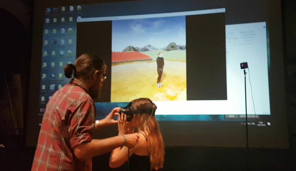 Two individuals interacting with virtual reality (VR) equipment. One person is assisting the other in putting on a VR headset. In the background, there is a large screen displaying a desktop with various icons and a window showing a virtual environment. The virtual environment appears to be an outdoor scene with a person standing on a yellow surface, with mountains and a blue sky in the background. There is also a VR sensor or camera on a stand to the right of the screen.