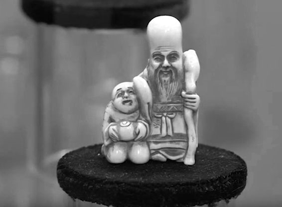A small figurine of two human-like figures. The larger figure is standing and holding a staff, while the smaller figure is kneeling and holding an object resembling a bowl or fruit. The figurine appears to be made of a light-colored material, possibly ivory or ceramic, and is placed on a dark circular platform. The background is blurred, emphasizing the figurine itself.