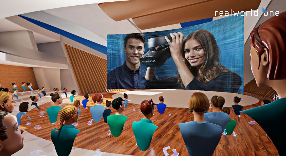 A virtual or augmented reality environment where a group of avatars is seated in an auditorium-like setting. The avatars are facing a large screen that displays an image of two people, one of whom is holding a virtual reality headset. The setting appears to be a virtual conference or presentation. The text 'realworld one' is visible in the upper right corner of the image. The auditorium has a modern design with wooden floors and curved seating arrangements. The avatars are diverse in appearance, suggesting a varied audience.